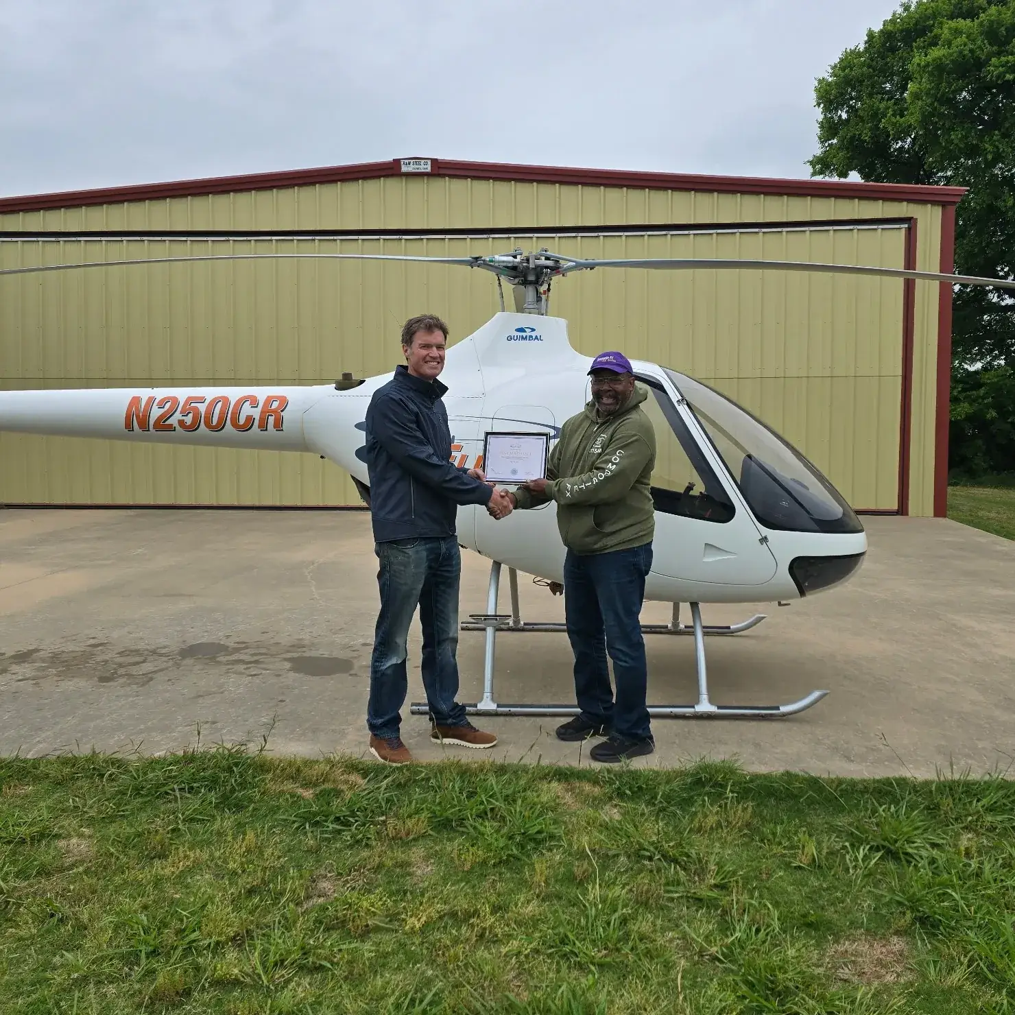 Student and instructor shaking hands and holding certificate in front of helicopter
